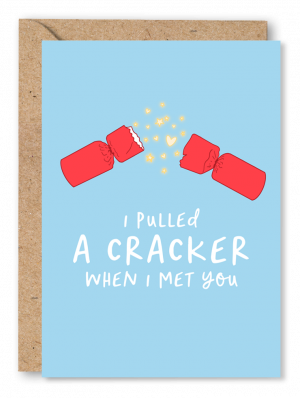 A blue Christmas card featuring an illustration of a pulled crack with hearts and stars exploding from the middle. White text below reads ‘I pulled a cracker when I met you’